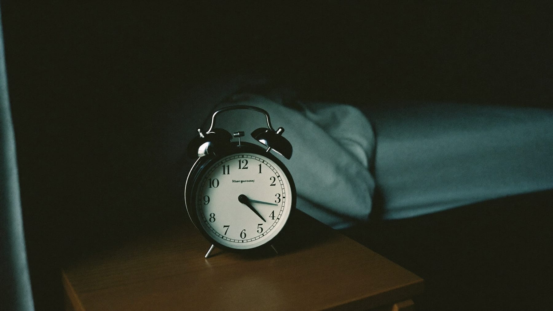 Despertador analógico numa mesa de cabeceira, num quarto às escuras, com a cama desfeita e vazia por trás