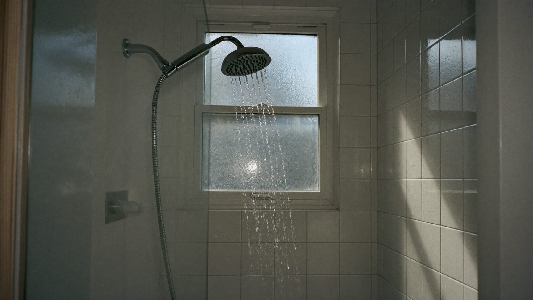 Água a correr de um chuveiro num duche de azulejos brancos, com luz fria da manhã a entrar por uma janela