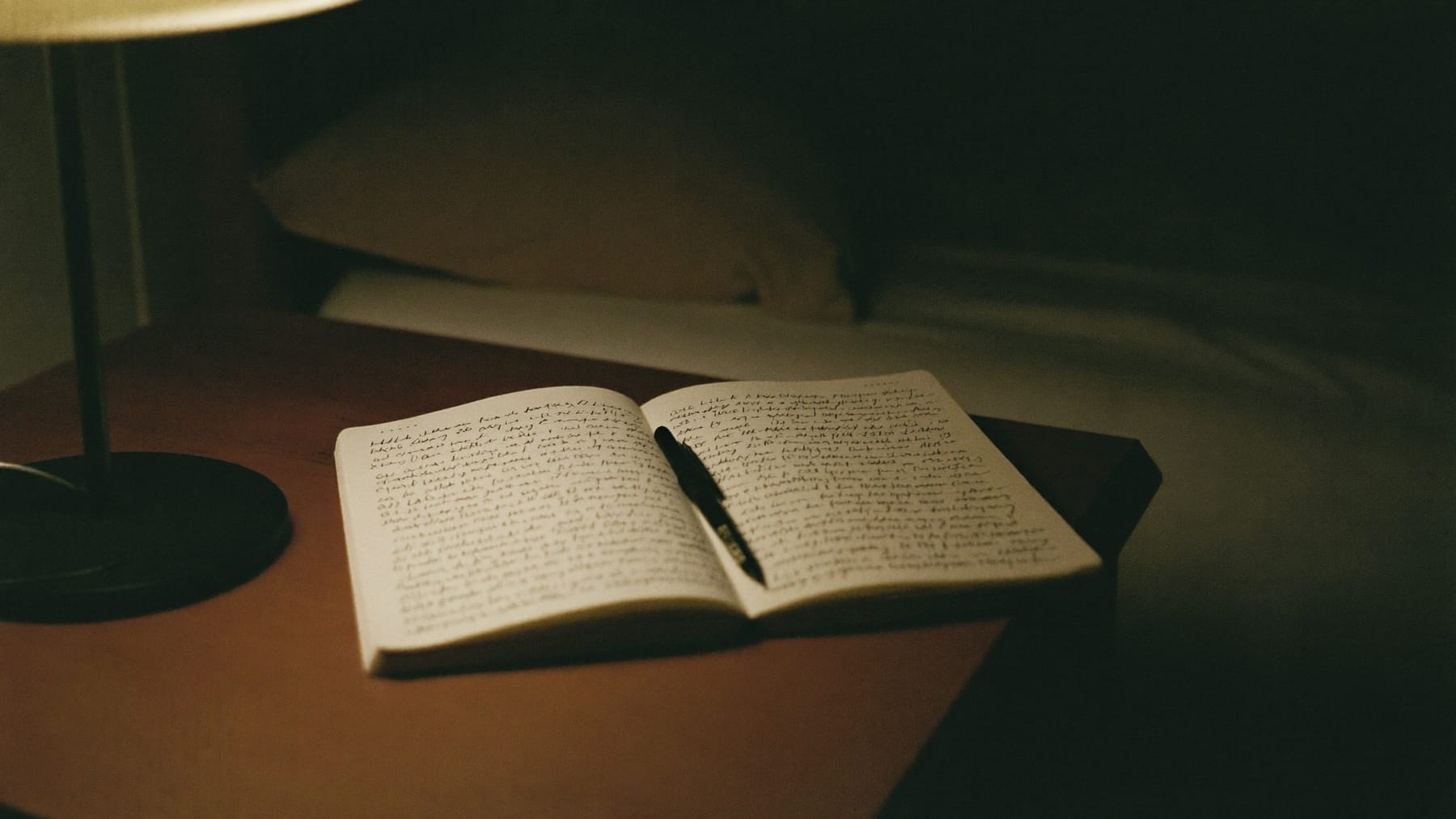 Caderno aberto com uma caneta pousada em cima, numa mesa de cabeceira iluminada por um candeeiro, com a cama por fazer em segundo plano