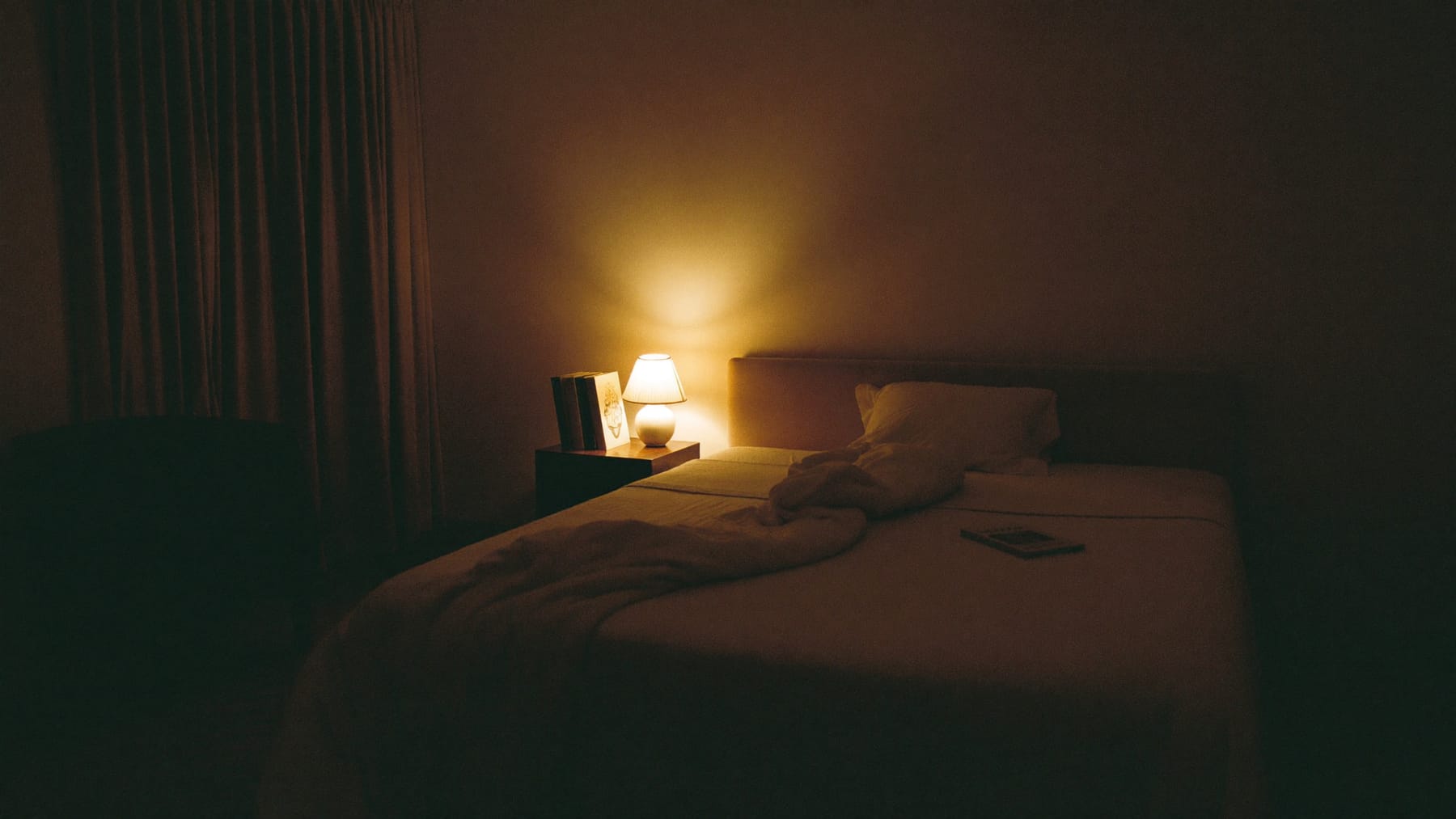 Quarto ao fim do dia, com a cama aberta e um candeeiro de cabeceira a dar a única luz