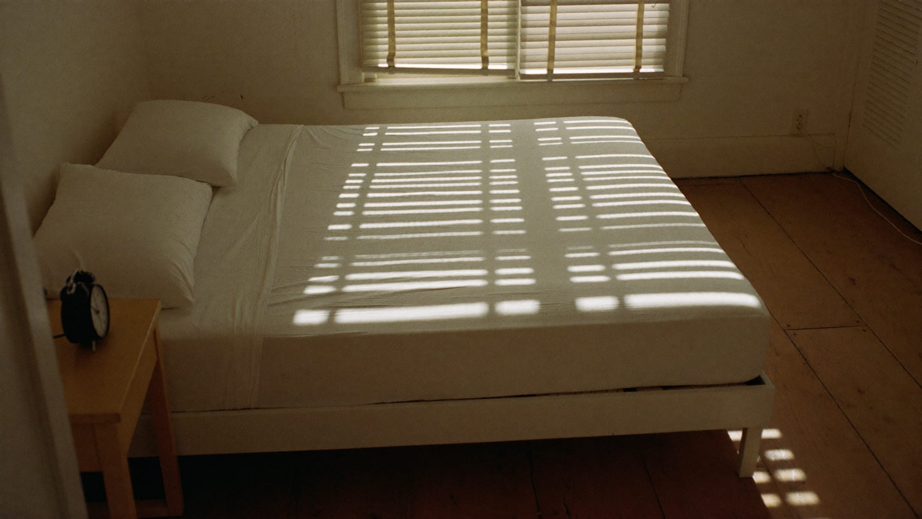 Cama por fazer com a luz do sol filtrada por estores a riscar os lençóis, e um despertador na mesa de cabeceira
