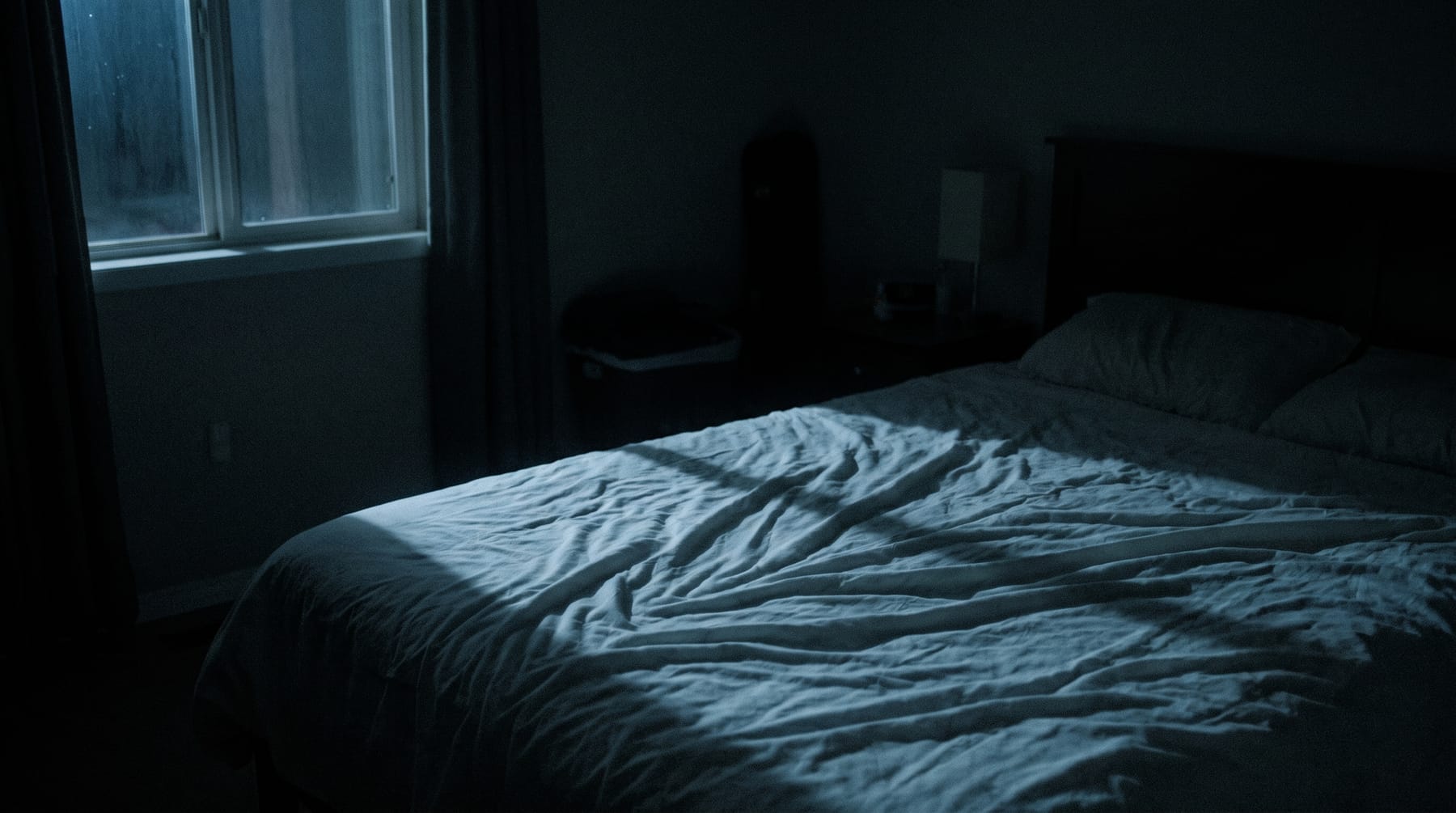 Quarto às escuras a meio da noite, com a luz fria da janela a cair sobre os lençóis desfeitos